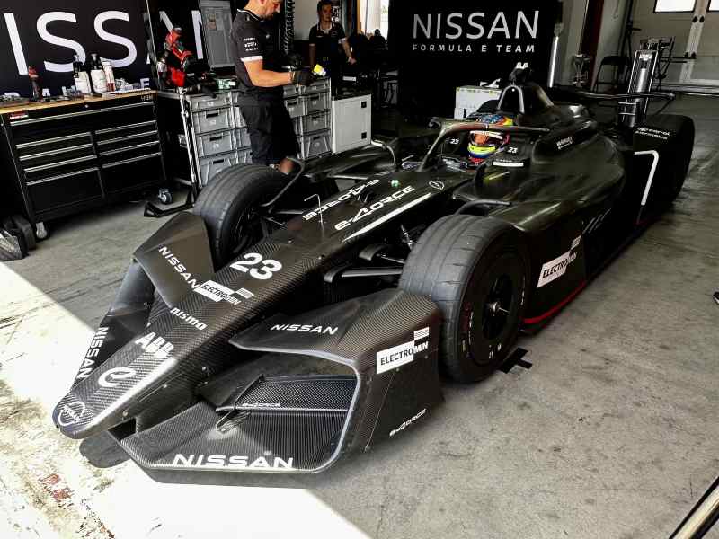 La fotografía muestra el monoplaza Nissan Formula E número 23 estacionado dentro del garaje del equipo. El vehículo luce una carrocería de fibra de carbono en acabado negro, con los logotipos de Nissan, NISMO y Formula E visibles en distintas partes del auto. Al fondo se observa a integrantes del equipo técnico trabajando en los preparativos previos a una sesión en pista. La imagen destaca la tecnología, aerodinámica y diseño característicos de la categoría eléctrica de monoplazas.