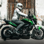 Motociclista con equipo de protección conduce una motocicleta de turismo urbano frente a una fachada tradicional de piedra y madera.