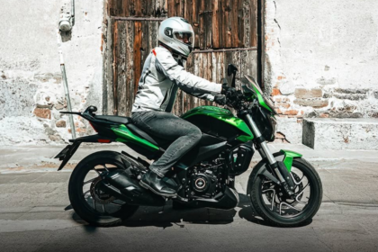 Motociclista con equipo de protección conduce una motocicleta de turismo urbano frente a una fachada tradicional de piedra y madera.
