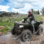 Vehículo todoterreno Polaris Sportsman 850 cruzando un arroyo en una ruta off road rodeada de montañas y vegetación natural.