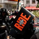 Asistente sostiene un banderín con la palabra RIDE y el logotipo de Harley-Davidson mientras un grupo de motociclistas participa en el European H.O.G. Rally 2026.