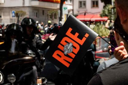 Asistente sostiene un banderín con la palabra RIDE y el logotipo de Harley-Davidson mientras un grupo de motociclistas participa en el European H.O.G. Rally 2026.