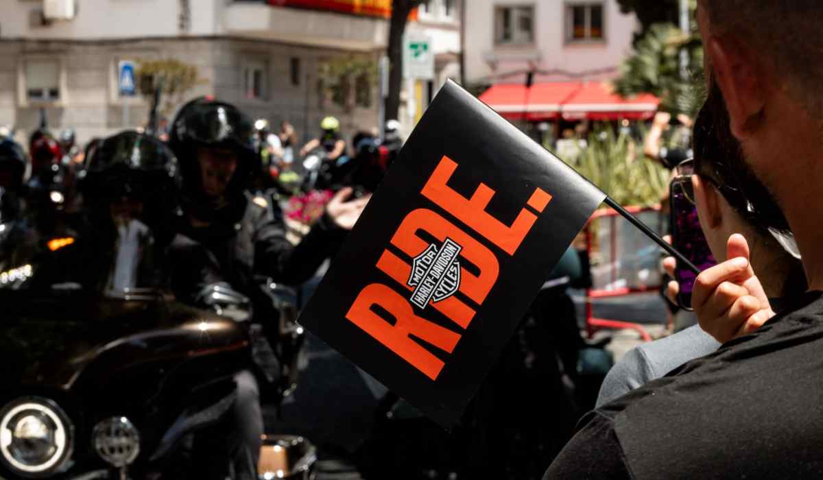 Asistente sostiene un banderín con la palabra RIDE y el logotipo de Harley-Davidson mientras un grupo de motociclistas participa en el European H.O.G. Rally 2026.