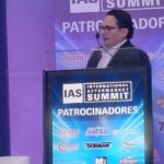 Ponente participa en una conferencia del International Aftermarket Summit 2026 desde el podio principal, frente a un escenario con logotipos de patrocinadores de la industria automotriz aftermarket.