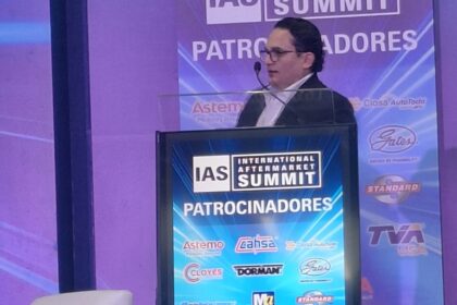Ponente participa en una conferencia del International Aftermarket Summit 2026 desde el podio principal, frente a un escenario con logotipos de patrocinadores de la industria automotriz aftermarket.