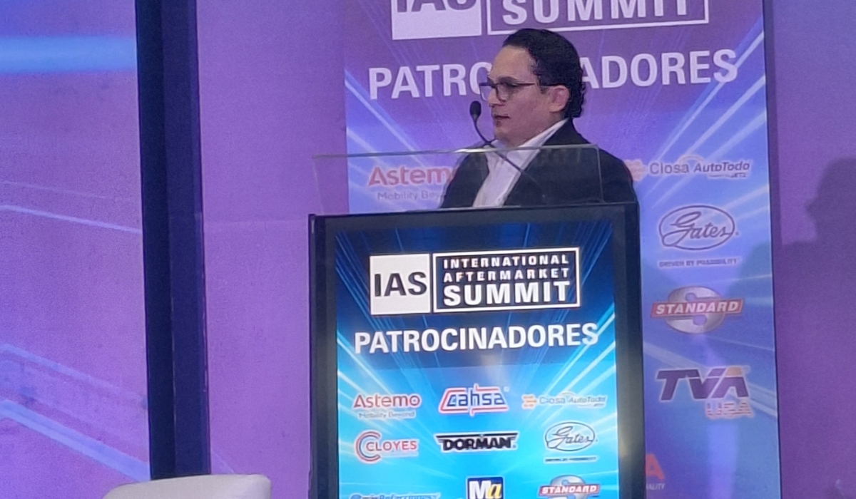 Ponente participa en una conferencia del International Aftermarket Summit 2026 desde el podio principal, frente a un escenario con logotipos de patrocinadores de la industria automotriz aftermarket.