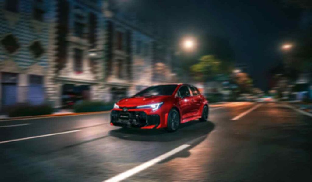 Toyota GR Corolla 2026 en color rojo circula de noche por una avenida urbana, destacando su diseño deportivo y tecnología desarrollada por GAZOO Racing.