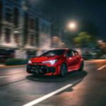 Toyota GR Corolla 2026 en color rojo circula de noche por una avenida urbana, destacando su diseño deportivo y tecnología desarrollada por GAZOO Racing.