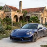 Porsche 911 azul oscuro circulando por un camino de terracería frente a una hacienda histórica henequenera durante el Rally de las Haciendas en Yucatán.