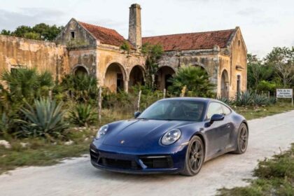 Porsche 911 azul oscuro circulando por un camino de terracería frente a una hacienda histórica henequenera durante el Rally de las Haciendas en Yucatán.