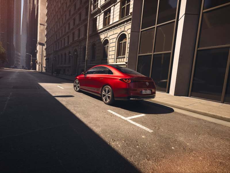 Vista trasera tres cuartos del nuevo Mercedes-Benz CLA eléctrico rojo en una calle urbana flanqueada por edificios.