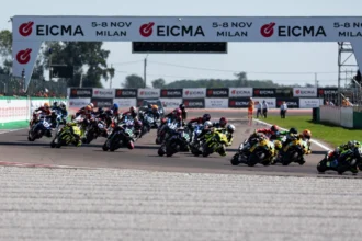 Grupo de motociclistas compitiendo en la pista bajo un arco publicitario del campeonato WorldSBK en el Circuito de Cremona.