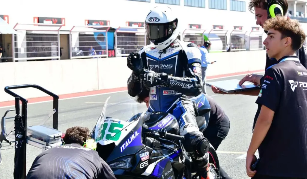 Piloto de ITALIKA Racing con indumentaria azul y blanca ajustando su postura sobre la motocicleta en el pit lane junto al equipo técnico.