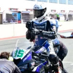 Piloto de ITALIKA Racing con indumentaria azul y blanca ajustando su postura sobre la motocicleta en el pit lane junto al equipo técnico.