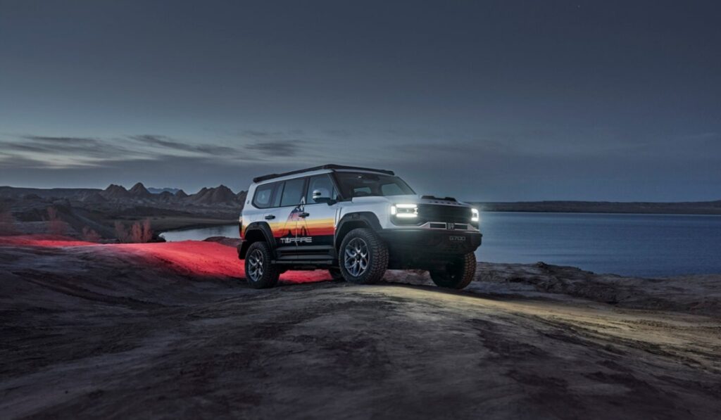 Camioneta todoterreno JETOUR G700 TOPFIRE de color blanco con vinilos laterales, faros encendidos y neumáticos todoterreno en un paisaje desértico junto al agua al atardecer.