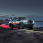 Camioneta todoterreno JETOUR G700 TOPFIRE de color blanco con vinilos laterales, faros encendidos y neumáticos todoterreno en un paisaje desértico junto al agua al atardecer.
