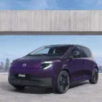 Vista frontal del coche urbano totalmente eléctrico Nissan PIXO en color morado oscuro estacionado frente a un paisaje urbano.