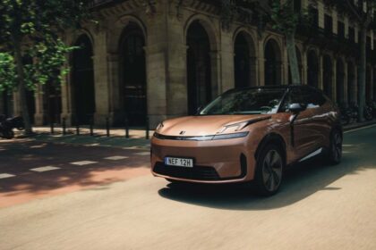Vehículo eléctrico Lynk & Co en tono cobre circulando por una calle urbana europea arbolada con arquitectura clásica.