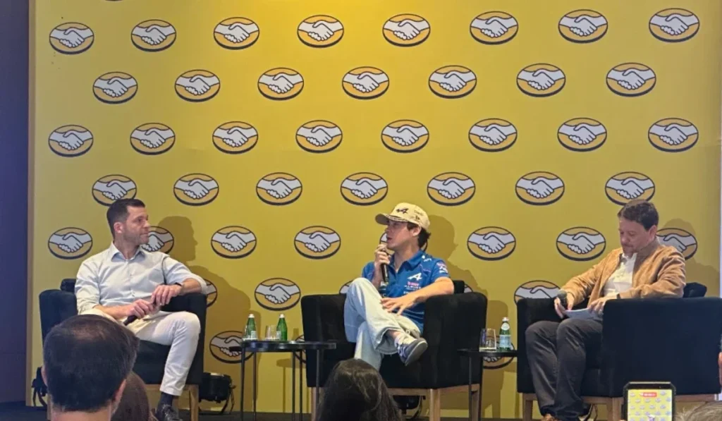 Franco Colapinto con gorra y uniforme de Alpine en un panel junto a David Geisen y Juan Lavista de Mercado Libre ante un fondo amarillo con logotipos de la marca.